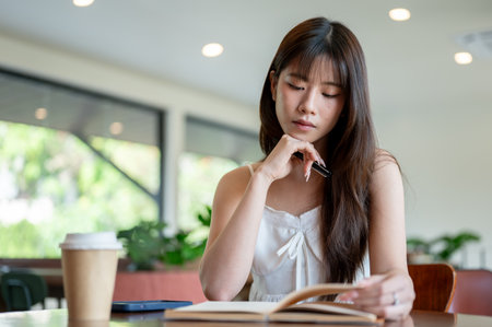 Asian woman in camisole holding a pen and put a hand under chin while reading a book with a coffee cup on cafe's table.の写真素材