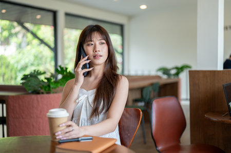 Asian woman in camisole is talking on the phone and grabbing her coffee while sitting at wooden table in the cafe.の写真素材