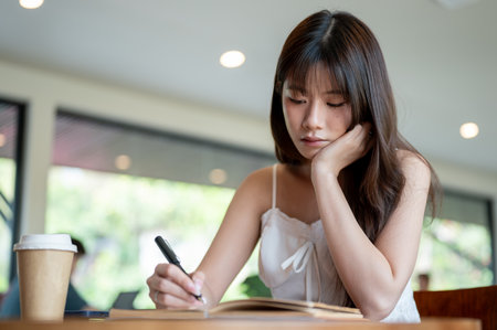 Asian woman in camisole with a hand under chin holding a black pen writing in notebook while sitting with coffee on table. Study or Working Outside.の写真素材