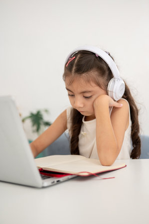 Asian kid little girl is looking at notebook with head resting on one hand while sitting at white table in house. Doing Homework or Online class.の写真素材