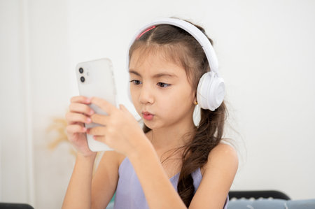 Asian kid or a girl wearing headphones holding phone with both hands while sitting at table in the house. Watching video or listening to music.の写真素材