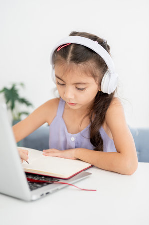 Asian kid little girl wearings white headphones and looking at a laptop on table while sitting in the house. Watching videos or listening to music.の写真素材
