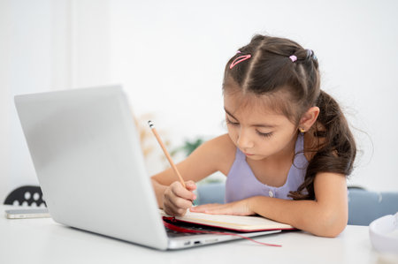 Asian kid little girl looking down at an open notebook over a laptop while sitting at white table in the house. Doing Homework or Online lesson.の写真素材