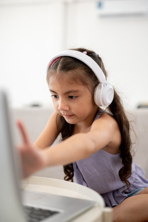 A kid or girl wearing headphones touching on her laptop screen at white table while sitting on sofa. Watching video or listening to music.の写真素材