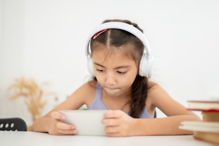 Asian kid or girl wearing headphones holding phone horizontally playing game with both hands while sitting at table. Watching video or listening to music.の写真素材