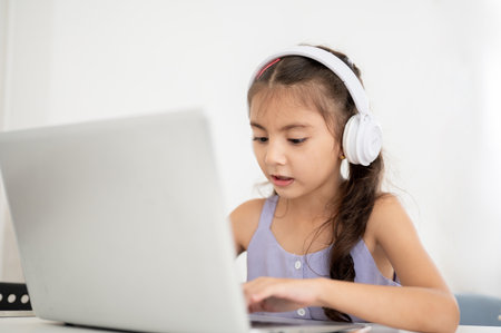 Asian kid or girl wearing headphones looking at laptop with open mouth while sitting at table in house. Searching videos or chatting with a friend.の写真素材