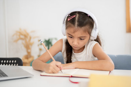 Asian kid little girl wearing headphones and holding pencil writing in notebook while sitting at the white table in house. Learning or Doing Homework.の写真素材