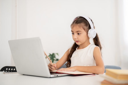 Asian kid little girl wearing headphones is writing on her notebook while looking at laptop on white table. Watching Video or Having an online class.の写真素材
