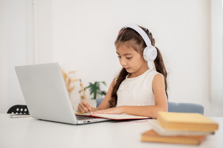 Asian kid little girl wearing headphones writing on her notebook while sitting with laptop on white table. Watching Video or Having an online class.の写真素材