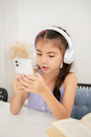 Asian kid or girl with headphones looking at phone in both hands with mouth open while sitting at table in house. Watching video or listening to music.の写真素材