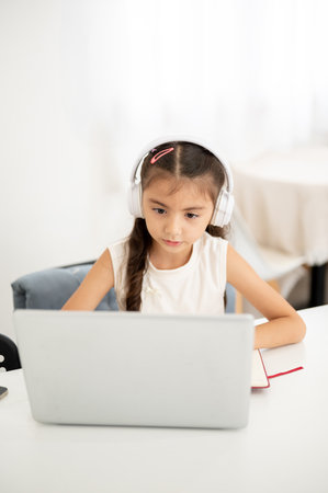 Asian kid little girl wearing headphone is looking intently at the laptop while sitting at white table. Watching Video or Having an online class.の写真素材