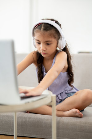 Asian kid or girl in purple shirt wearing headphone is typing on her laptop at white table while sitting on sofa. Watching video or listening to musicの写真素材
