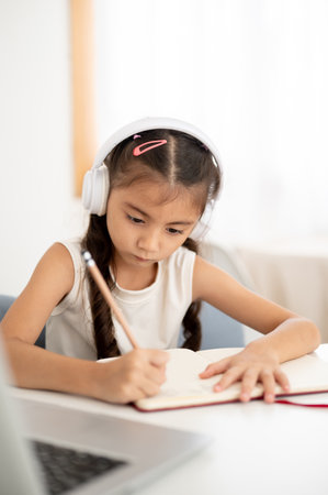 Asian kid little girl wearing headphones and writing in notebook with a pencil while sitting at white table in the house. Learning or Doing Homework.の写真素材