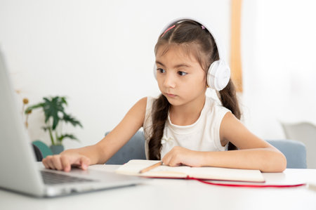 Asian kid little girl wearing headphones and looking at her laptop while sitting at white table in the house. Learning or Doing Homework.の写真素材