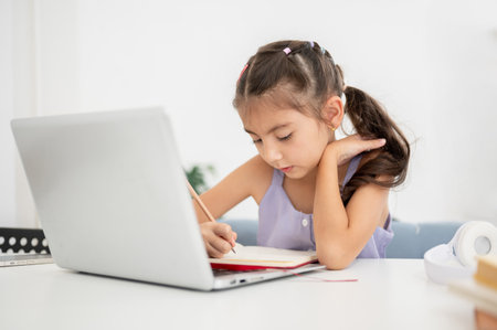 Asian kid little girl writing in notebook over laptop with hand touching hair while sitting at white table in house. Doing Homework or Online lesson.の写真素材