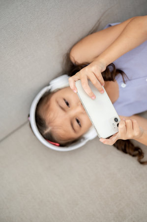 Asian kid or little girl wearing white headphones holding smartphone with both hands while lying on fabric sofa. Watching video or listening to music.の写真素材