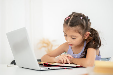 Asian kid little girl reading or looking at an open notebook over a laptop while sitting at white table in the house. Doing Homework or Online lesson.の写真素材