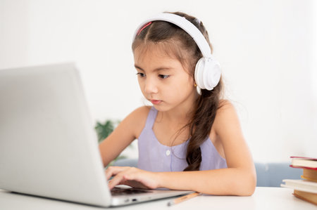 Asian kid or a girl wearing headphones is staring and typing on laptop while sitting at table in house. Searching videos or chatting with a friend.の写真素材