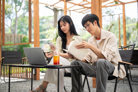 Asian man wearing glasses is reading a notebook while a woman looking at phone at folding table....