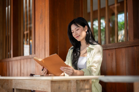 Long hair asian woman is smiling and reading a book while sitting at table in a wooden house. Working or Study outdoor.の写真素材