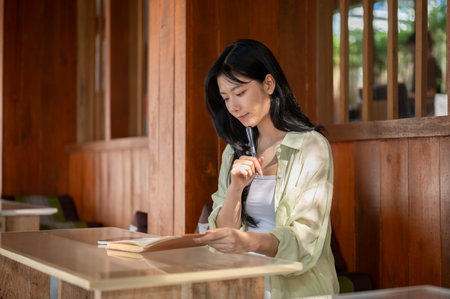 Asian woman holding pen under chin and looking down at a book while sitting in wooden house. Working or Study outdoor, Traditional Cafe or Restaurant.の写真素材
