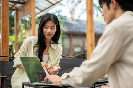 Smiling asian woman leaning to look at her friend's laptop screen while sitting on folding chair. Working or Study outdoor, Garden Cafe or Restaurant.の写真素材