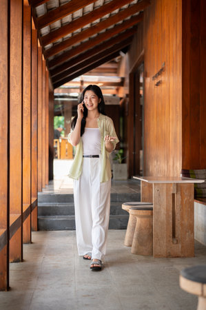 Long hair asian woman is smiling during a phone call while walking in a wooden house's corridor. Chilling outdoor, Traditional Cafe or Restaurant.の写真素材