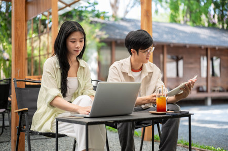 Asian woman looking at her laptop while man reading a notebook sitting together at folding table....