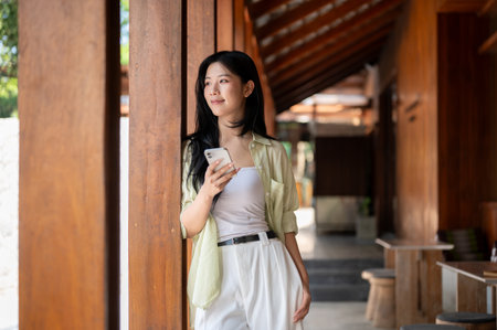 Long hair asian woman standing and leaning on a pillar while holding phone in wooden house corridor. Chilling outdoor, Traditional Cafe or Restaurant.の写真素材