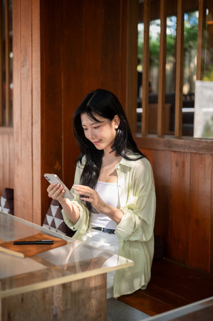 Asian woman is smiling and looking at smartphone in hand while sitting at table in a wooden house. Chilling outdoor, Traditional Cafe or Restaurant.の写真素材