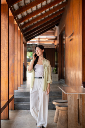 Long hair asian woman talking on a smartphone while standing or walking in wooden house's corridor. Chilling outdoor, Traditional Cafe or Restaurant.の写真素材