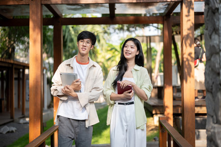 Smiling asian friends or a man and woman holding books and standing together in wooden corridor....