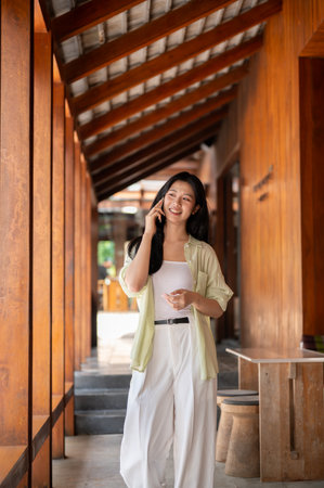 Long hair asian woman is standing and talking on a smartphone in the wooden house's corridor. Chilling outdoor, Traditional Cafe or Restaurant.の写真素材