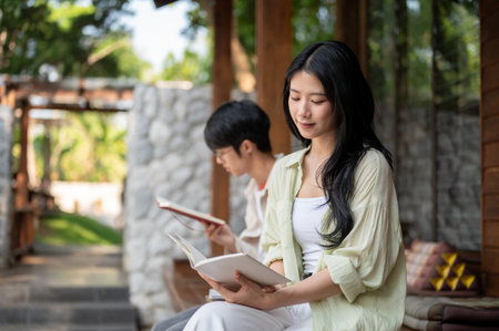 Long hair asian woman reading a white book while sitting cross legged next to a man at wooden terrace. Chilling Outdoor, Garden Cafe or Restaurant.の写真素材