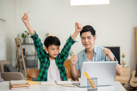 A son raising one arm and father making a fist while looking at laptop together in their house. Elementary Schoolboy, Kids education, Family bonding.の写真素材