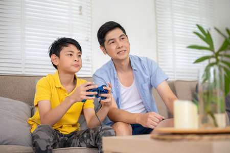 Asian father talking while elementary kid son holding joystick with both hand playing video game at sofa in their house. Fun activity, Family bonding.の写真素材