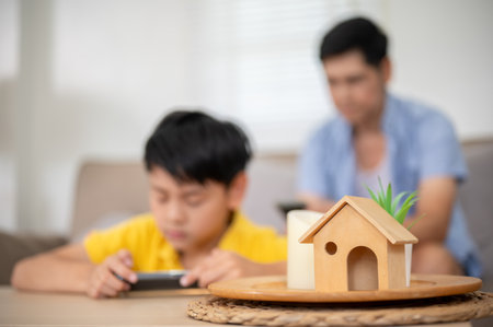 Close up of the wooden house toy on table with blurred background of father and son holding smartphone in living room. Kid block toys, Fun activity.の写真素材