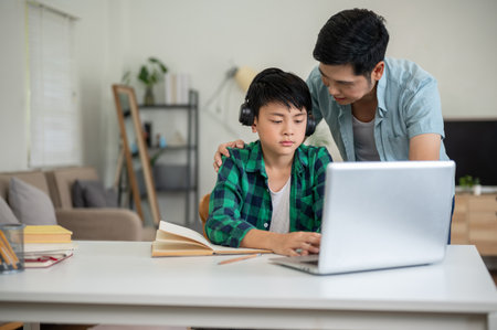 Asian kid elementary school boy wearing headphones looking at laptop on table while father is talking. Online Study, Doing homework, Internet surfing.の写真素材