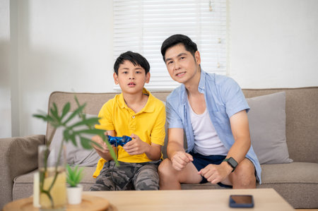 Asian father looking ahead as his elementary school kid son holding joystick playing video game at...