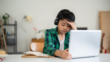 Asian kid elementary school boy wearing headphone looking at laptop on table with hand touching head. Online Study, Doing homework, Internet surfing.の写真素材