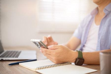 Side view close up of a man hand holding smartphone over notebook with laptop aside at wooden table in office or working room. Workspace Setup.の写真素材