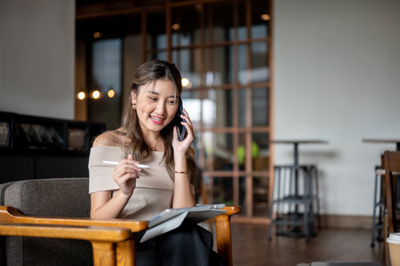 Asian woman talking on smartphone with a tablet on her lap while sitting on wooden chair in cafe or coffee shop. Chilling Outdoor, Working or Study.の写真素材