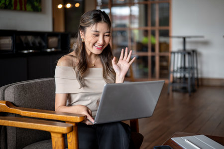 Asian woman waving a hand greeting on a video call via laptop while sitting in cafe or coffee shop. Working or Study. Online class or meeting.の写真素材
