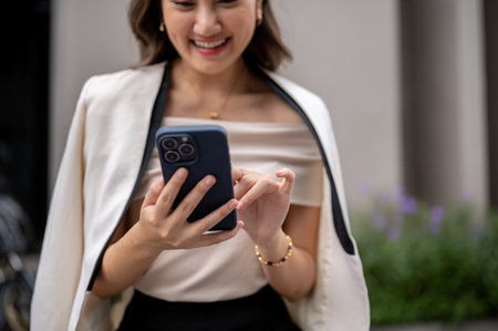 Close up of a long hair asian woman is holding and looking at her smartphone while walking around the building. Urban Life, Chilling Outdoor.の写真素材
