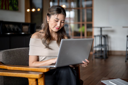 Asian woman sitting and placing laptop on her lap at wooden armchair in the cafe or coffee shop. Working or Study. Coworking space.の写真素材