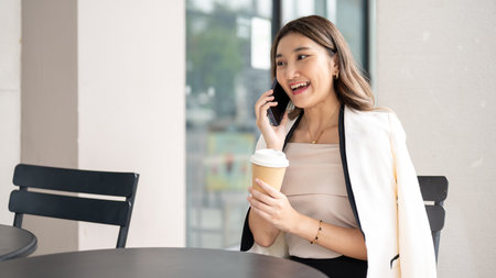 Long hair asian woman smiling during a phone call and holding a coffee cup while sitting at black table in the cafe. Urban Life, Chilling Outdoor.の写真素材