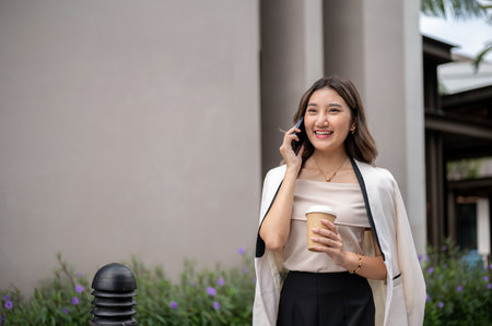 Long hair asian woman talking on the phone while walking on the path around the building with a coffee cup in her hand. Urban Life, Chilling Outdoor.の写真素材