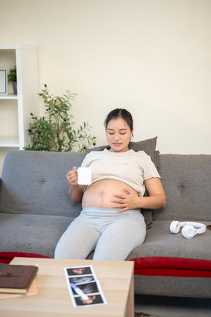Pregnant asian woman holding a cup drinking water and touching her bare belly while sitting on sofa...