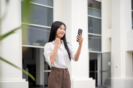Smiling asian woman in white shirt holding phone and making a fist while walking out of university or office building. Video call or recording herself.の写真素材