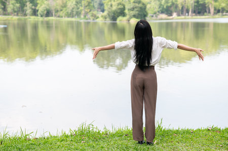 Back view of a woman in white shirt stretching or standing with open arms on grass at the side of lake or river. Relaxing Outdoor, Chilling in park.の写真素材